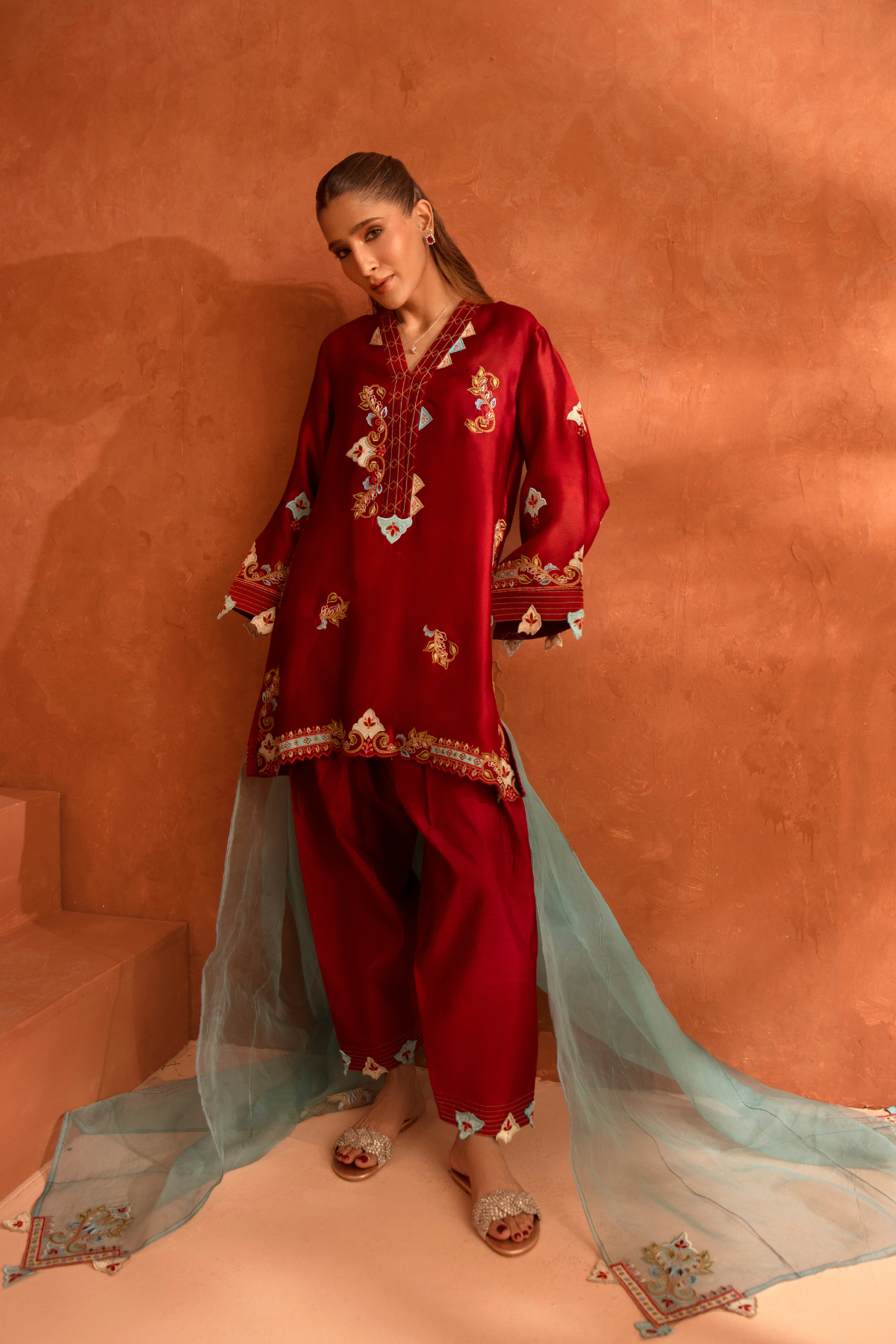 Woman wearing a red embroidered traditional outfit with a sheer dupatta against an orange wall.