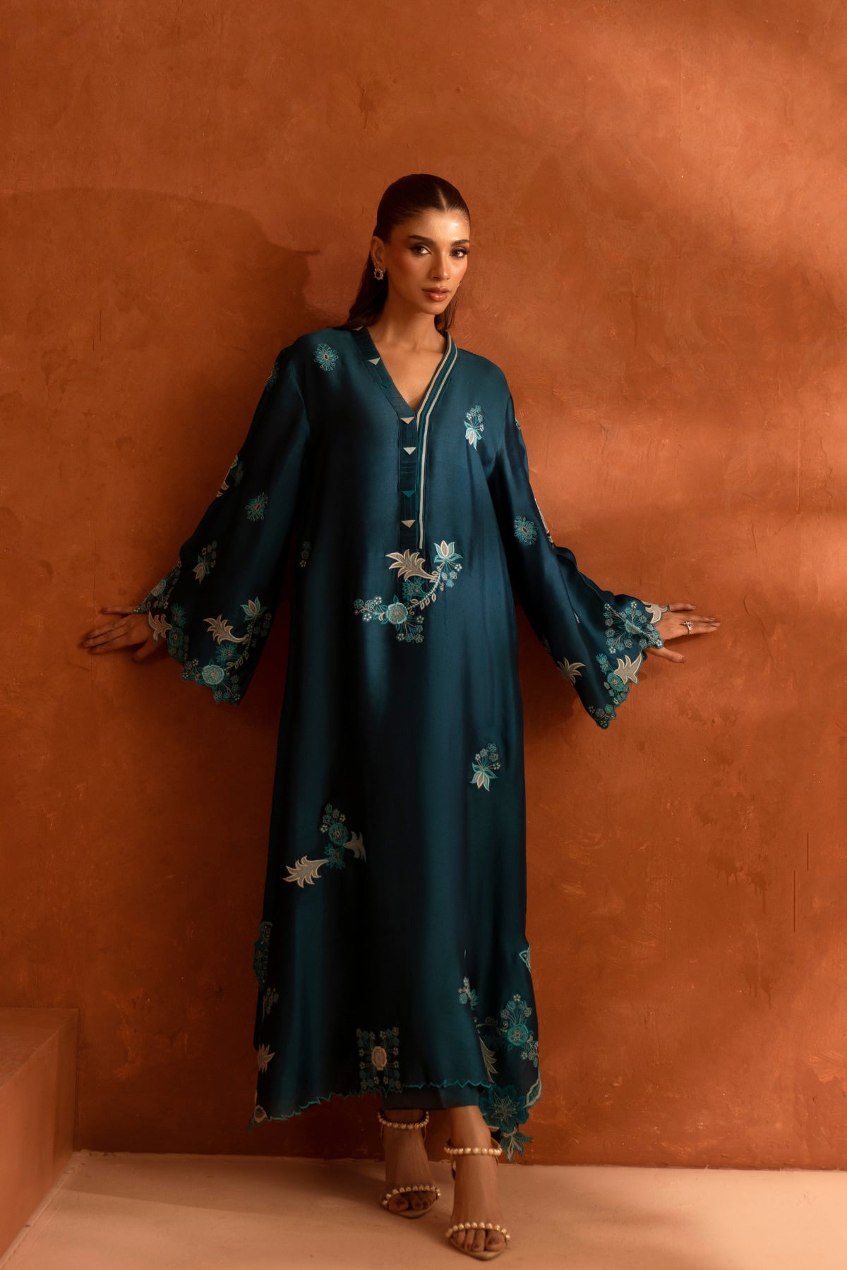 Woman wearing a teal dress with floral patterns against a brown wall.