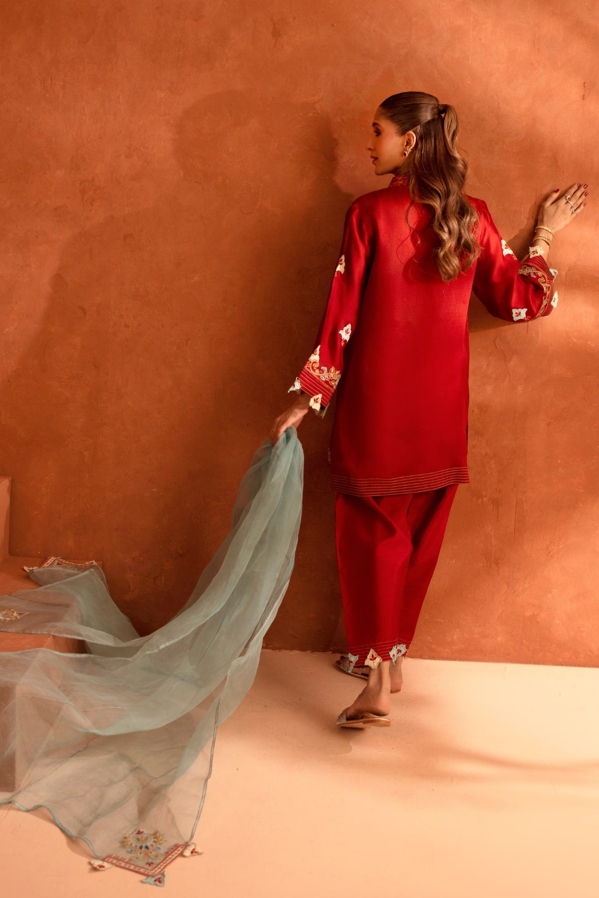 Woman in a red traditional outfit holding a light blue dupatta against a brown background